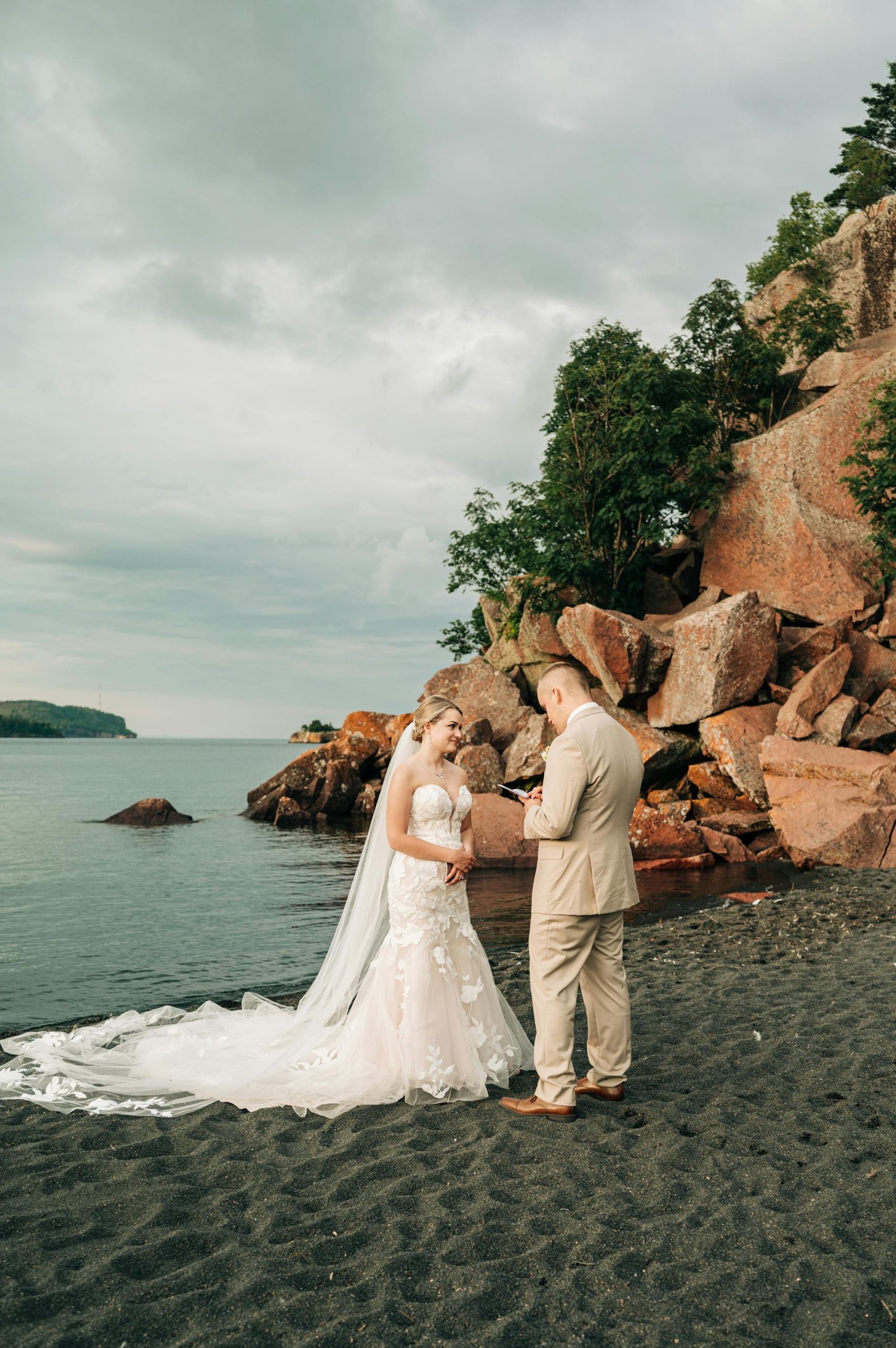 Bride wearing Hattie by Rebecca Ingram on a black sand beach listening to her groom read his wedding vows