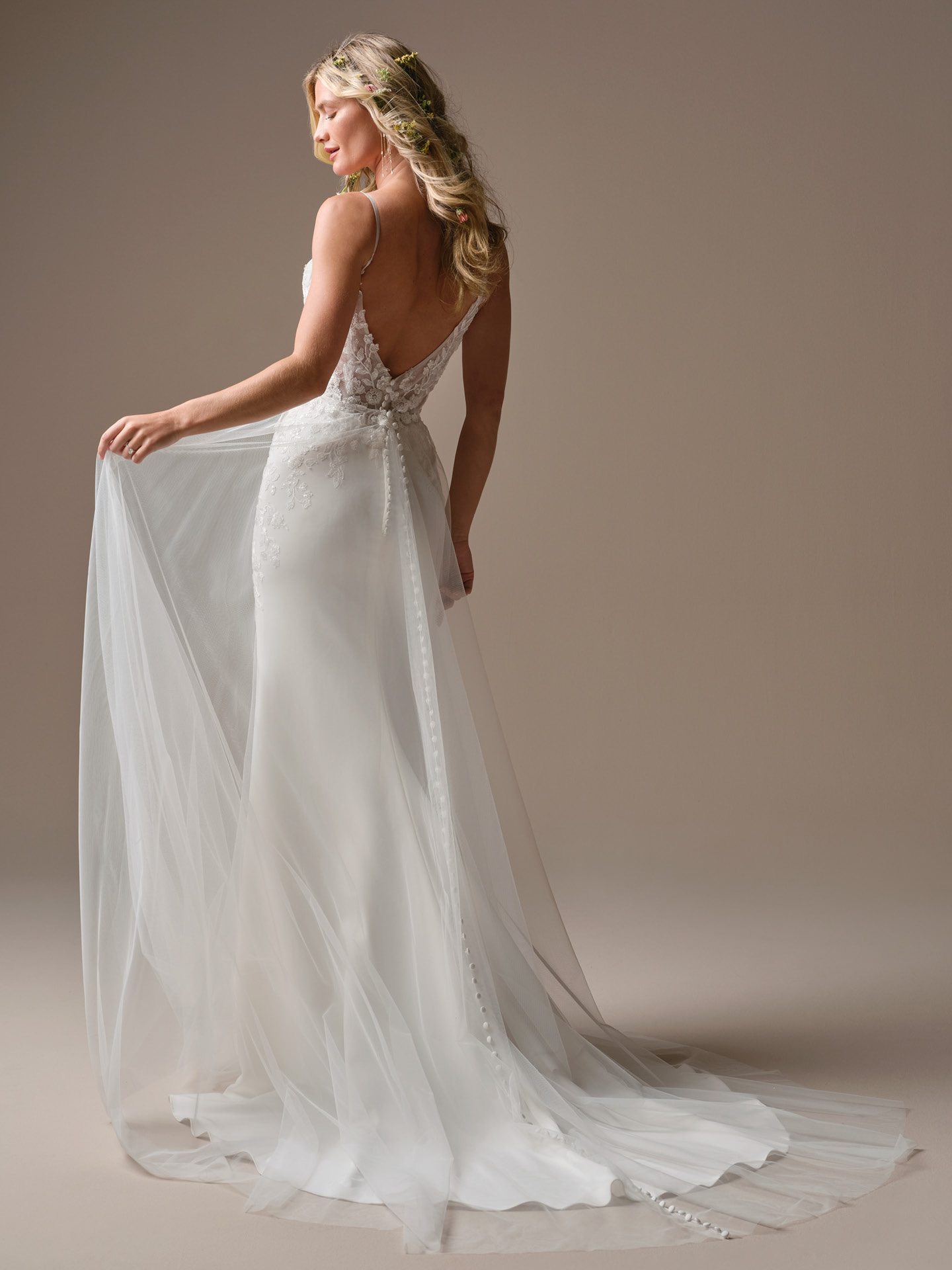 Back view of a fitted wedding gown with a removable tulle overskirt and delicate button detailing, creating a flowing train effect over a sleek silhouette.