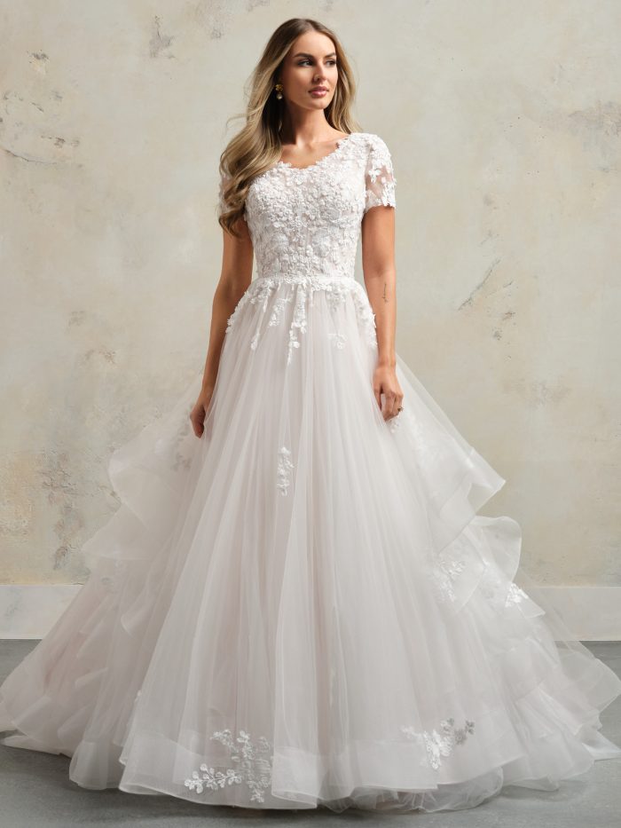Bride wearing Winona Leigh by Rebecca Ingram, a modest A-line wedding dress with pleated tulle bodice, lace motifs, short illusion lace sleeves, and layered skirt with horsehair hem ruffles.