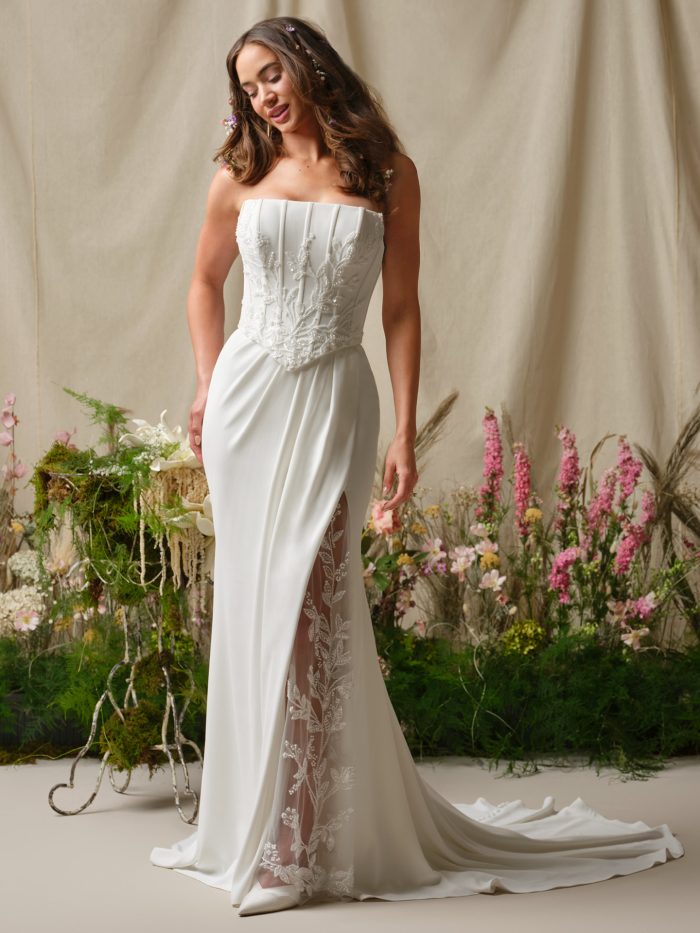 Strapless fitted wedding dress named Terry with a structured embroidered bodice, soft draping at the waist, and a sheer floral lace slit, shown on a model standing in a garden-inspired setting.