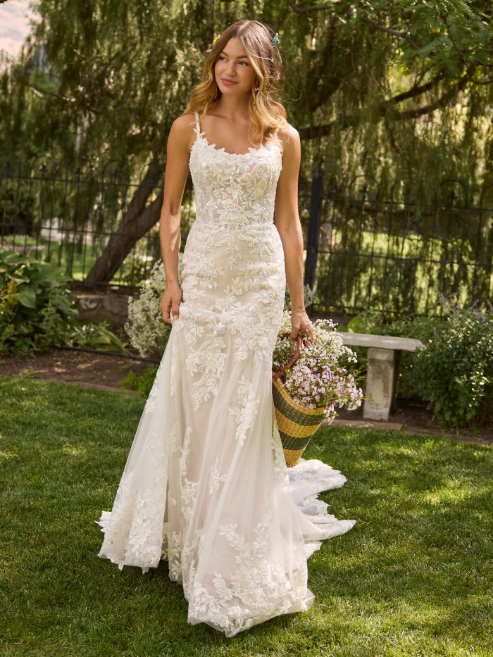 Bride wearing Rebecca Ingram Eden fit-and-flare wedding dress with delicate floral lace appliqués, spaghetti straps, and a soft illusion bodice, holding a basket of wildflowers in a garden setting.