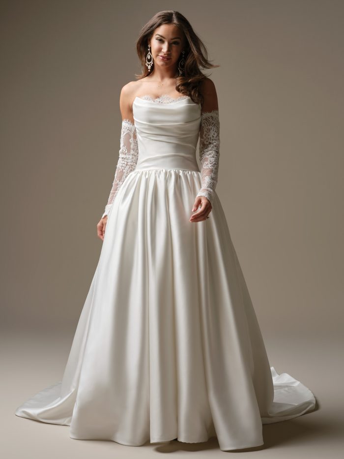 A model wearing the Maggie Sottero Sylvana wedding dress, featuring a draped satin bodice with lace trim, a full satin ball gown skirt and detachable lace arm sleeves.