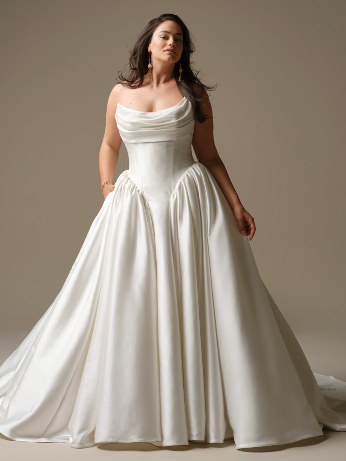 Curve model wearing Hudson by Maggie Sottero — strapless satin ballgown wedding dress with draped cowl neckline, exposed corset seams, and gathered pleats at the waist, styled with gold drop earrings and flowing hair.