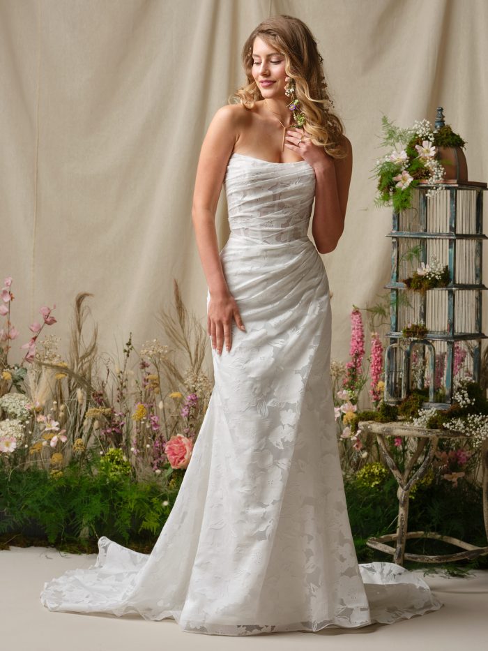 Bride wearing the Kendall sheath wedding dress featuring a strapless cat-eye neckline, printed organza fabric, luxe ruching, and a softly fitted silhouette with a subtle train, styled in a romantic garden-inspired studio setting.