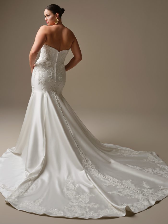 Back view of Burke by Sottero and Midgley — plus-size fit-and-flare satin wedding gown featuring chapel-length train with lace appliqué edging and covered button detailing down the back.
