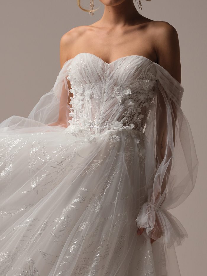 Close-up detail of the Ireland wedding dress and detachable sleeves by Maggie Sottero, showing 3D floral lace appliqué on the ruched sweetheart bodice with sheer draped off-the-shoulder tulle sleeves gathered at the wrist.