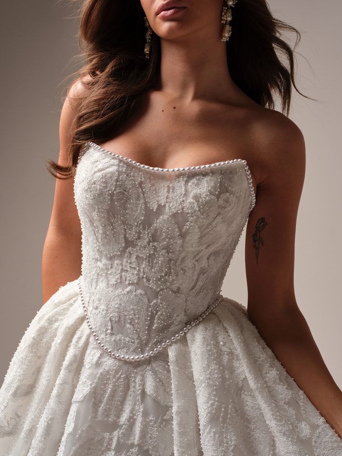 Close-up detail of the January wedding dress bodice by Sottero and Midgley, showing the strapless sweetheart neckline with pearl beading, intricate floral lace appliqué, and a beaded belt cinching the waist.