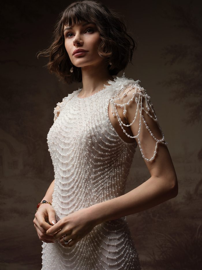 Close-up detail of the Oleta sheath wedding dress and cap sleeves by Sottero and Midgley, showing scalloped pearl beading over the bodice with a floral shoulder accent and draped pearl chains cascading down the arm.