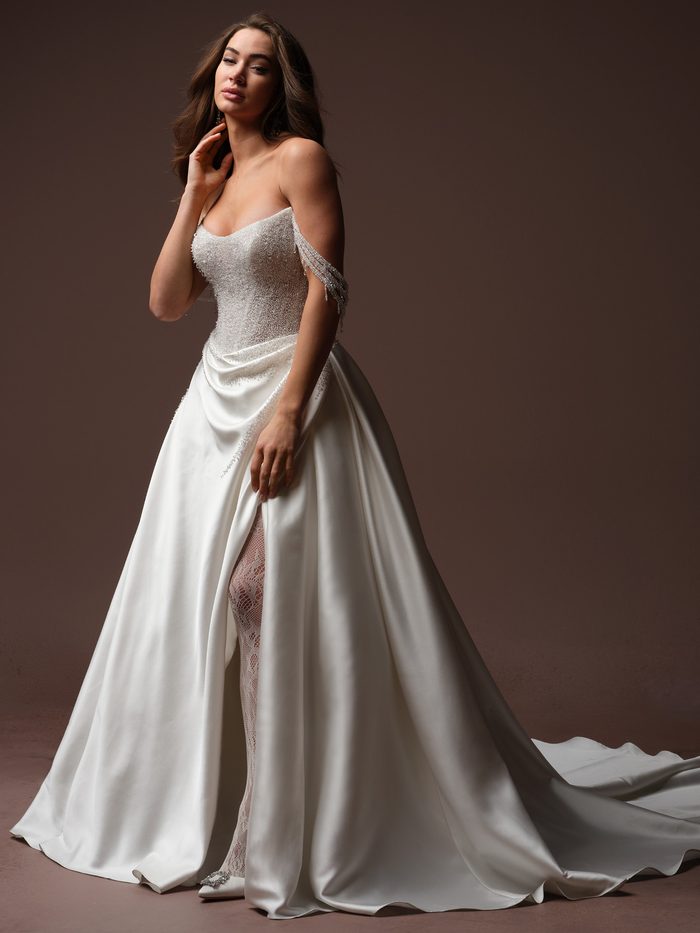 Full-length side view of the Rumi ball gown wedding dress by Sottero and Midgley, featuring a beaded sweetheart bodice with cascading crystal fringe straps, a draped satin ball gown skirt with a front slit revealing a lace underlay, and a chapel train.