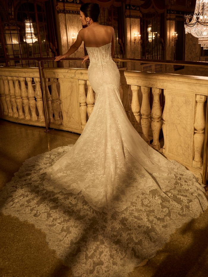 Maggie Sottero Bellevue Royale fit-and-flare wedding dress featuring a strapless lace bodice with covered buttons down the back and a dramatic lace train, shown from the back on an ornate balcony.