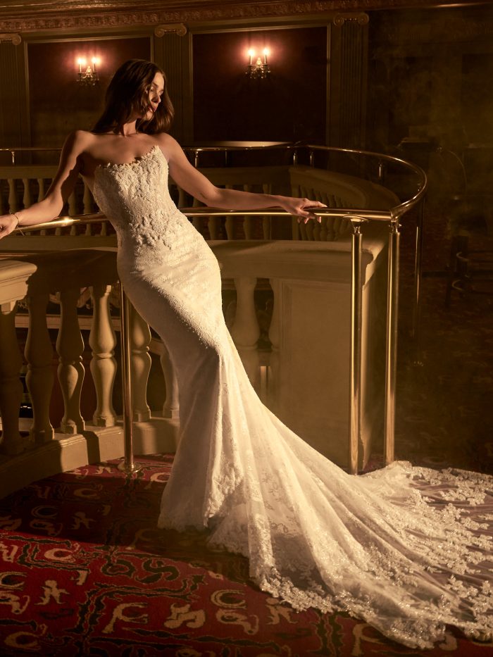 Maggie Sottero Collette sheath wedding dress with fitted lace silhouette and extended lace train, shown draped elegantly along a balcony railing in a richly detailed venue.