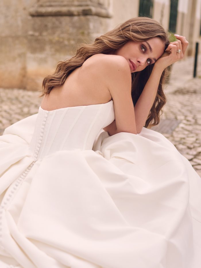Bride seated in a strapless satin ball gown wedding dress with covered button back detailing and full skirt, resting her head on her hand with soft, romantic expression.