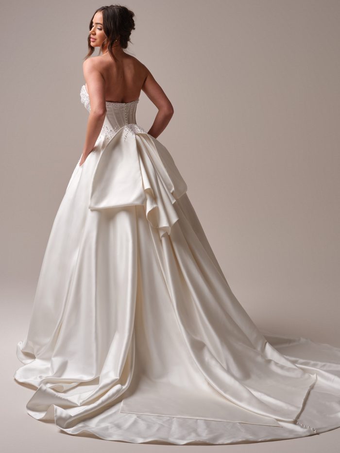 Back view of a strapless satin ball gown wedding dress with a beaded corset bodice and oversized bow detail at the waist, featuring a long dramatic train.