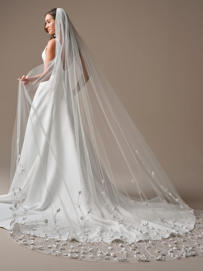 Bride wearing the Lawrence veil by Maggie Sottero, featuring a cathedral-length design with 3D floral and pearl embellishments cascading along the hem.