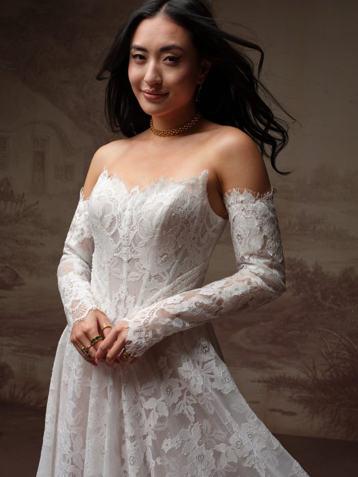Maggie Sottero Mischa A-line wedding dress in all-over floral lace with a sweetheart neckline trimmed in eyelash lace, and detachable sheer lace off-the-shoulder long sleeves with matching eyelash lace cuffs.