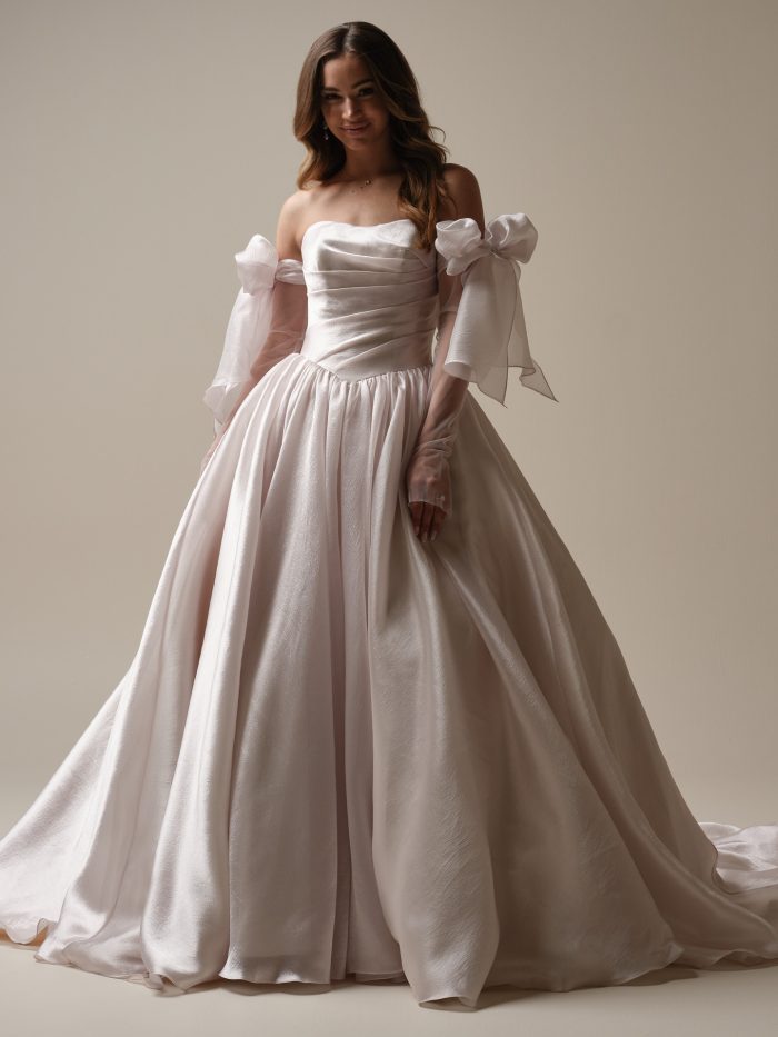 Bride wearing a strapless satin ball gown wedding dress with ruched bodice and detachable off-the-shoulder organza sleeves tied in bows, softly lit against a neutral background.