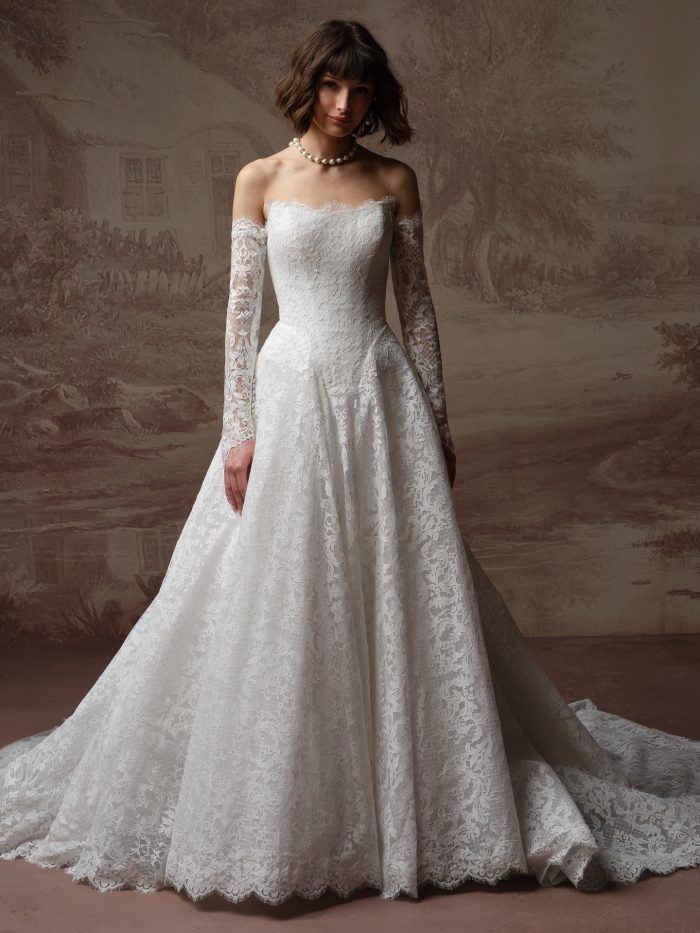 Maggie Sottero Serena A-line wedding dress in all-over lace with a straight scalloped neckline, detachable sheer lace off-the-shoulder long sleeves with an ornate damask lace pattern, and a full cathedral train.
