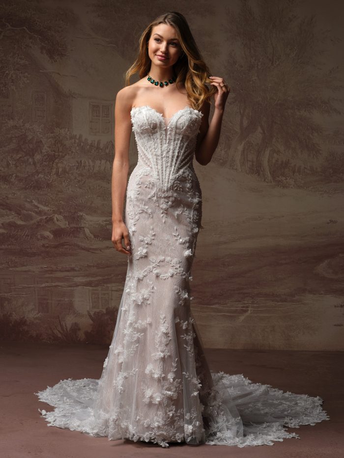 Maggie Sottero Stasia fit-and-flare wedding dress with a strapless sweetheart neckline, textured floral appliqués, and a fitted silhouette that flares into a delicate lace train, photographed in a studio setting.