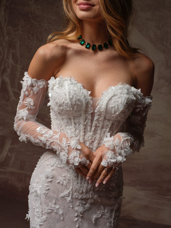 Stasia by Maggie Sottero fit-and-flare wedding dress featuring a sweetheart neckline, intricate 3D floral appliqués, sequin and beaded lace, and sheer off-the-shoulder long sleeves, styled with an emerald green gemstone choker necklace.