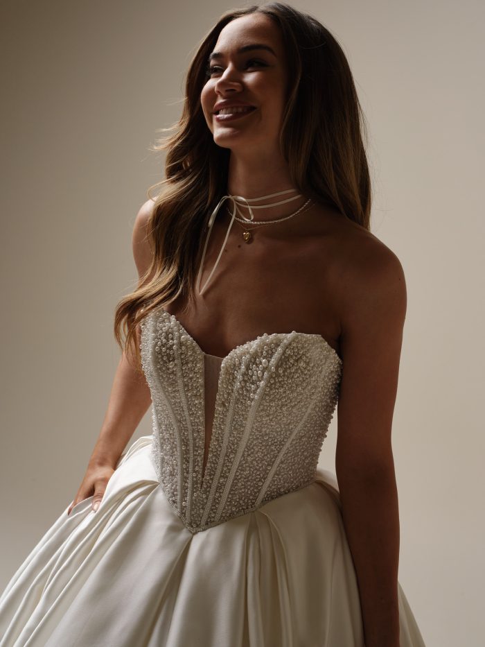 Close-up of a strapless satin ball gown wedding dress featuring a pearl-encrusted corset bodice with illusion plunge neckline and visible boning details, bride smiling while holding the skirt.