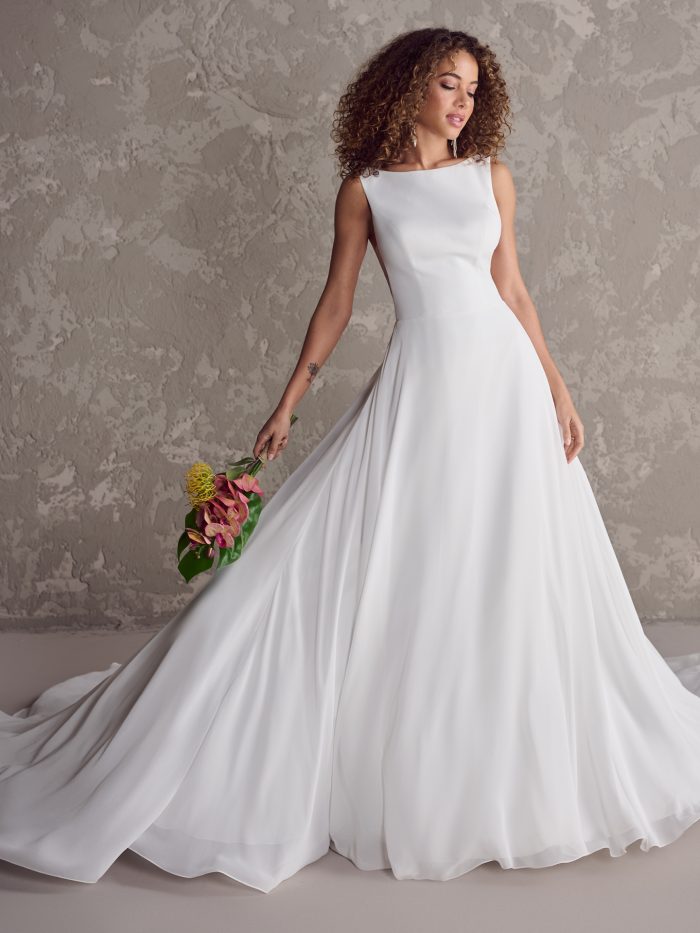 Bride wearing a sleeveless chiffon ball gown wedding dress with a bateau neckline and flowing skirt, holding a bouquet and posing against a textured gray backdrop.