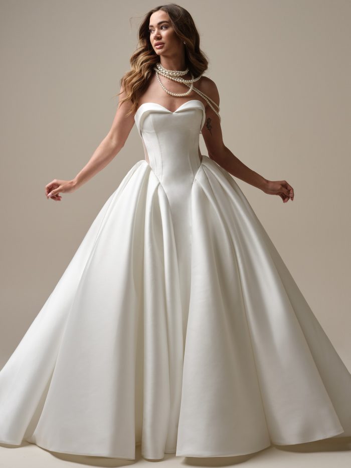 Bride wearing a strapless satin ball gown wedding dress with a sweetheart neckline, corset-style bodice, and side illusion cutouts, styled with layered pearl accessories.