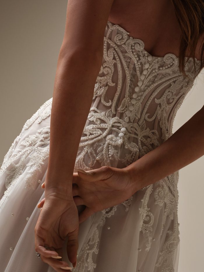 Close-up of an off-the-shoulder beaded lace A-line wedding dress showing the intricate embroidery, covered button back, and pearl detailing on the sheer tulle fabric.