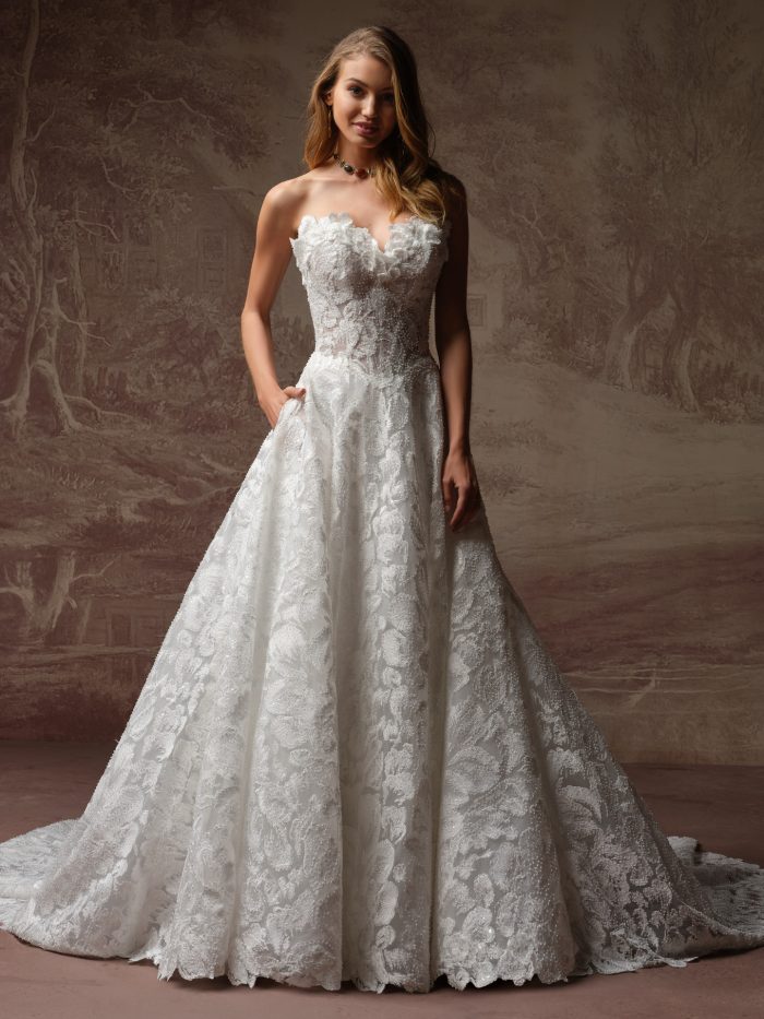 Sottero and Midgley Merrigan A-line wedding dress with an off-the-shoulder lace bodice and full skirt with intricate floral texture, photographed full-length against a softly painted backdrop.