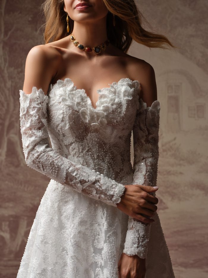 Sottero and Midgley Merrigan A-line wedding dress close-up featuring an off-the-shoulder neckline with dimensional floral lace and sheer long sleeves, detailed with textured appliqués and beadwork.