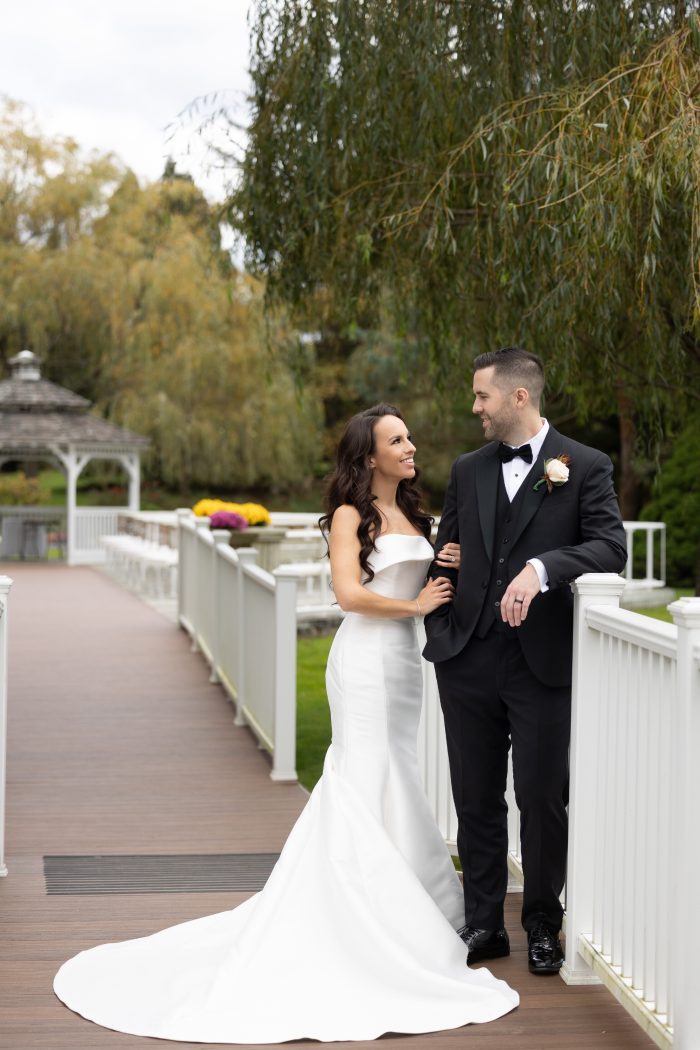 Bride and groom smile at each other while standing on a white bridge in a garden setting. The bride wears a sleek strapless mermaid wedding dress with a long train, and the groom wears a black tuxedo with a boutonniere.