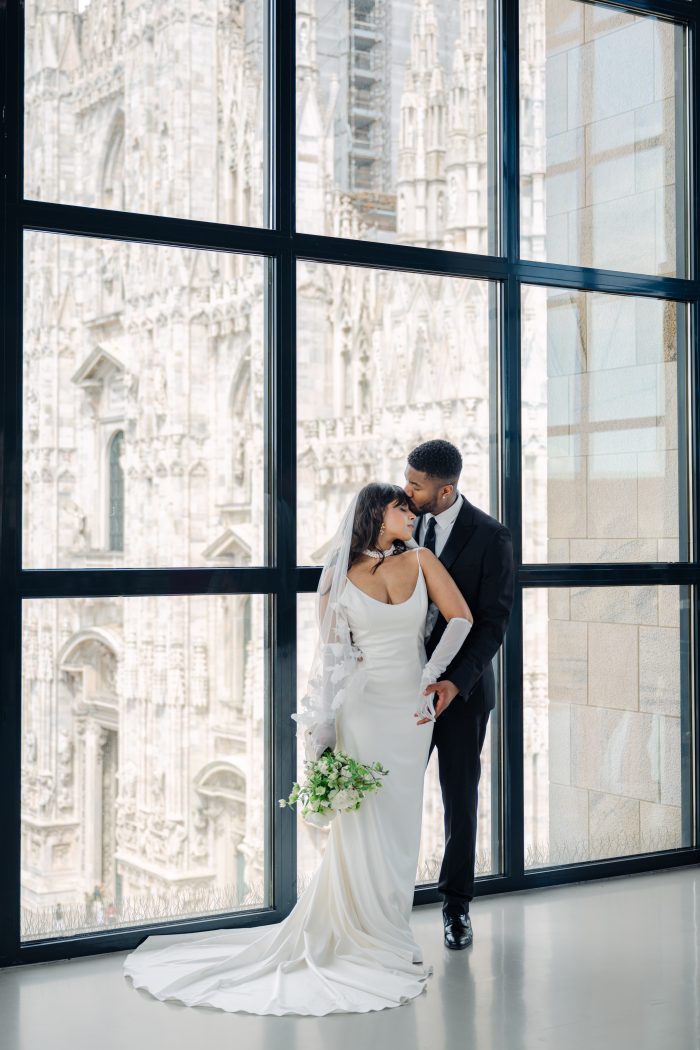 Bride and groom share a romantic moment indoors with the intricate facade of a European cathedral visible through tall windows. The bride wears a sleek satin gown with spaghetti straps, matching gloves, and a floral-embroidered veil, holding a bouquet of white and green flowers. The groom wears a classic black suit and tie.