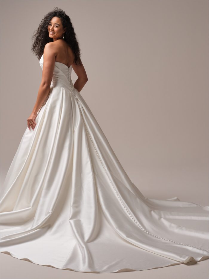 Bride in a strapless satin ballgown wedding dress with a ruched bodice and full pleated skirt, featuring fabric-covered buttons trailing down the train.