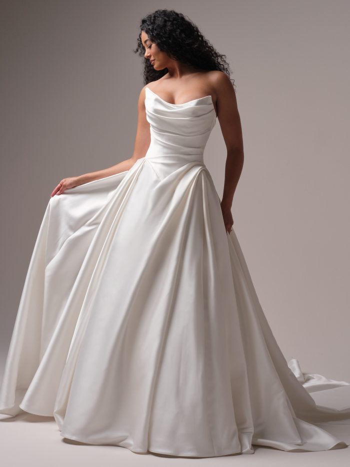Bride wearing a strapless satin ballgown wedding dress with a romantic cowl neckline and draped bodice, holding the side of her voluminous skirt.