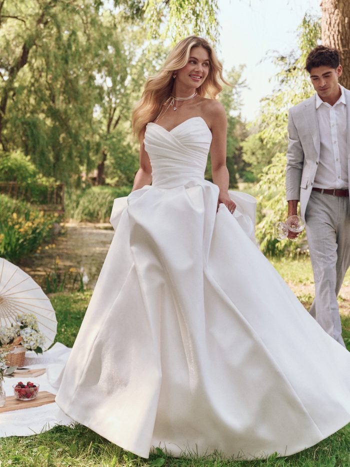 Bride in Maggie Sottero Aubrielle strapless ballgown wedding dress with a sweetheart neckline and structured pleats, walking through a romantic garden picnic scene.