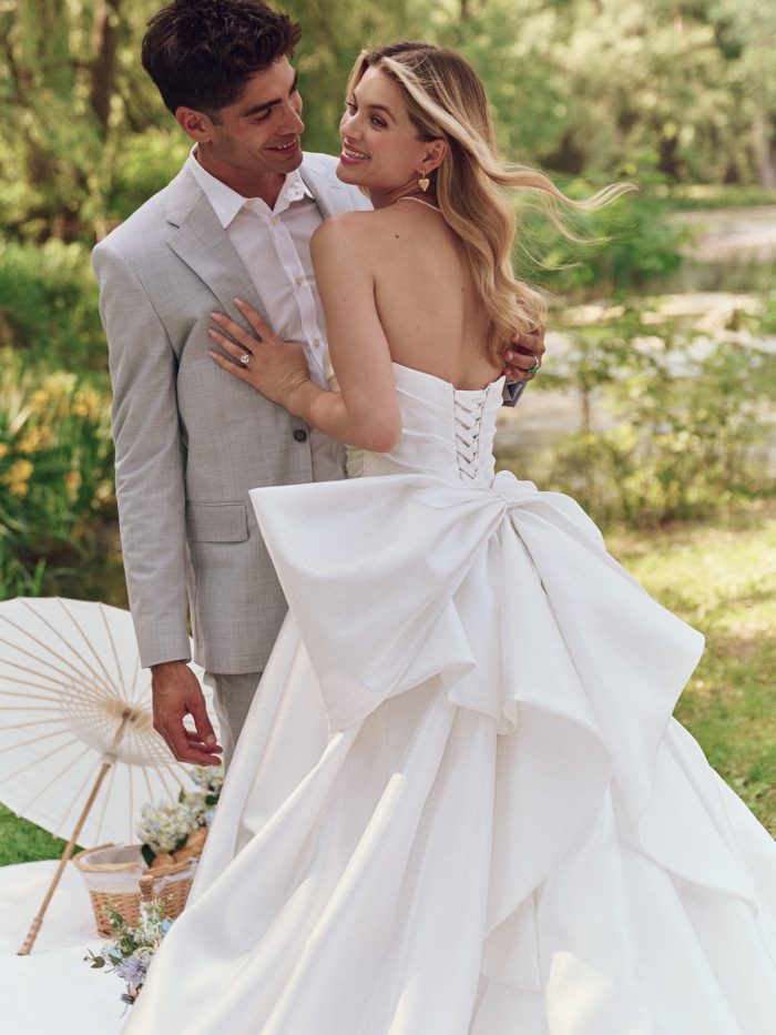 Bride wearing Maggie Sottero Aubrielle wedding dress, featuring a strapless pleated bodice, corset back, and dramatic oversized bow, embracing her partner in a garden setting.