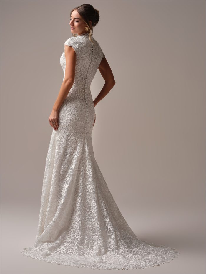 Back view of a modest sheath wedding dress with cap sleeves, a high neckline, and allover floral lace, featuring a scalloped train and covered button detailing.