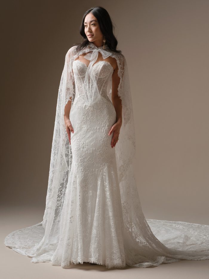 Bride wearing the Bellevue fit-and-flare wedding dress by Maggie Sottero, featuring a strapless lace corset bodice, floral lace detailing, and a dramatic sheer lace cape with an elegant train.
