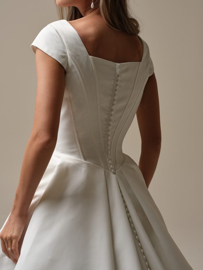Back view of a ballgown wedding dress with cap sleeves, square neckline, exposed vertical seams, and covered buttons trailing down the full pleated skirt.