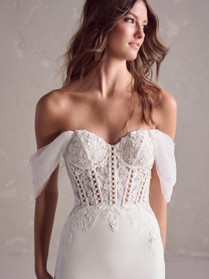 Bride wearing a strapless sweetheart wedding gown with a sheer lace bodice featuring visible boning, floral appliqués, and beaded embellishments. Delicate off-the-shoulder tulle sleeves drape over the arms, adorned with scattered pearls.