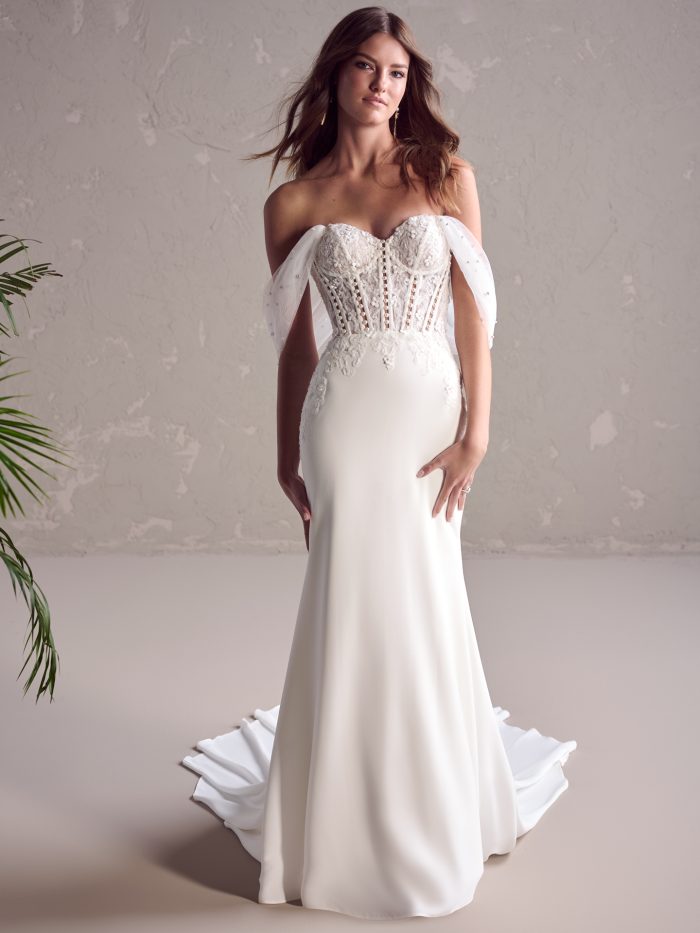 Bride wearing a fit-and-flare ivory wedding gown with a floral lace and illusion corset-style bodice, off-the-shoulder pearl-embellished tulle sleeves, and a sleek crepe skirt flowing into a short train.