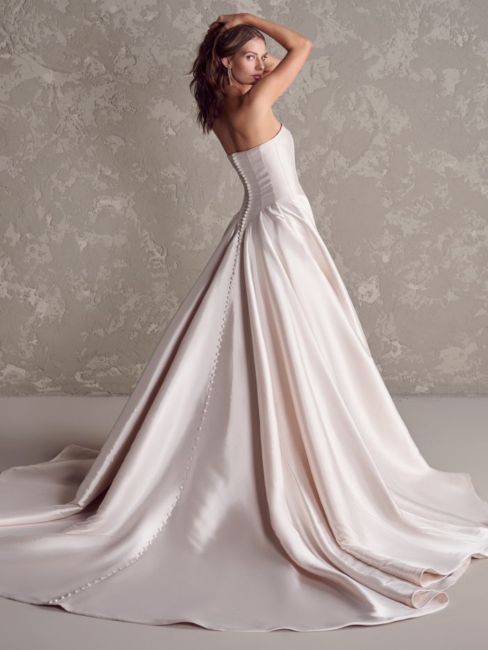 Back view of Maggie Sottero Emmanuelle satin ballgown wedding dress with a row of buttons trailing down the train, showcasing a flattering corset-inspired fit and elegant sweep train.