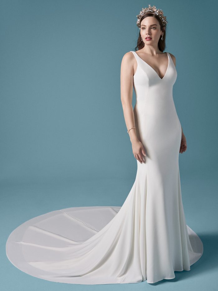 Front view of a minimalist sheath wedding dress with a deep V-neckline and wide straps, paired with a dramatic circular train and ornate tiara for a modern royal look.