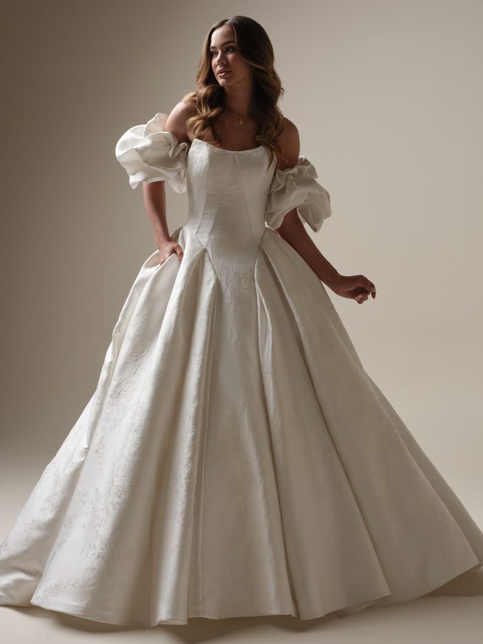 Bride wearing a romantic jacquard ballgown wedding dress with off-the-shoulder puff sleeves, a square neckline, and full pleated skirt with floral embossing and hidden pockets.