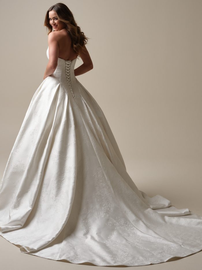 Back view of a strapless jacquard ballgown wedding dress with a lace-up corset closure and sweeping train, showcasing subtle floral texture and structured pleating.