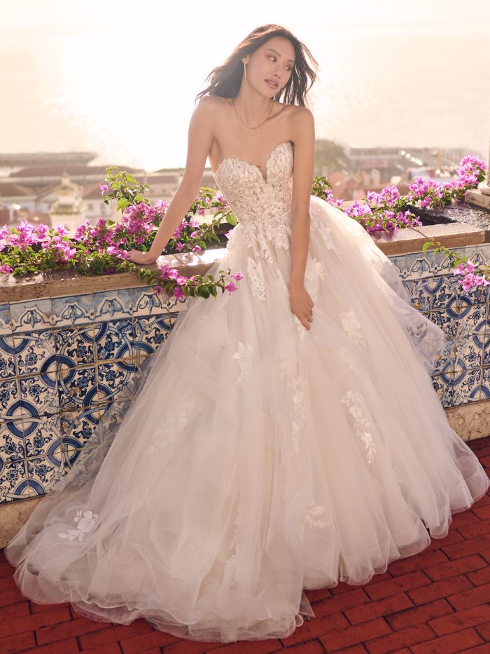 Bride wears Indiana by Maggie Sottero—a romantic strapless ballgown with floral lace bodice, sweetheart neckline, and voluminous tulle skirt, posing by a tiled terrace with ocean views.