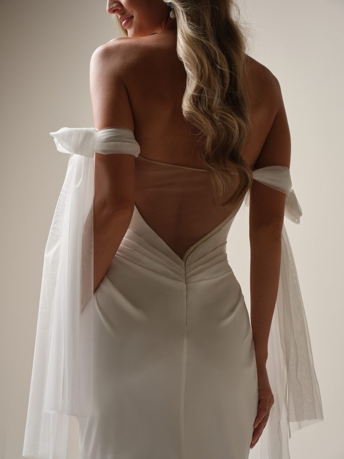 Back view of a bride wearing a ruched satin sheath wedding gown with sheer off-the-shoulder tulle sleeves and a low illusion back.