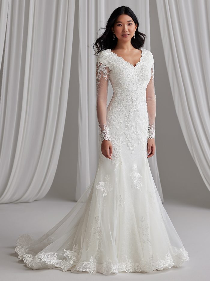Bride wearing a lace fit-and-flare wedding dress with illusion long sleeves, scalloped V-neckline, and floral motifs trailing into a soft tulle skirt.
