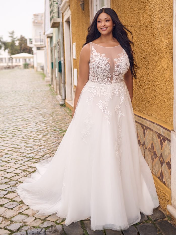 Front view of the Lindsey ball gown wedding dress, featuring an illusion bateau neckline, beaded lace bodice, and soft tulle skirt, worn by a smiling bride on a cobblestone street.