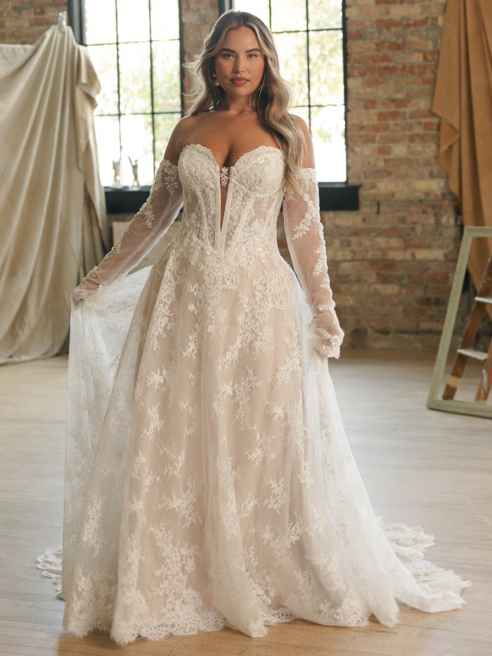Plus-size bride wearing the Lucca strapless lace A-line wedding dress with exposed corset boning, sheer off-the-shoulder long sleeves, and a soft tulle skirt in a sunlit studio.