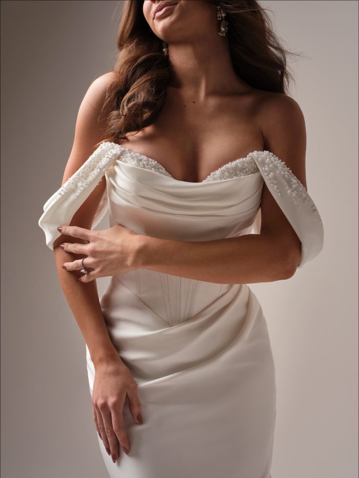 Close-up of a bride in a satin wedding dress with off-the-shoulder draped straps and pearl-beaded neckline, featuring a ruched bodice and visible corset-style structure.